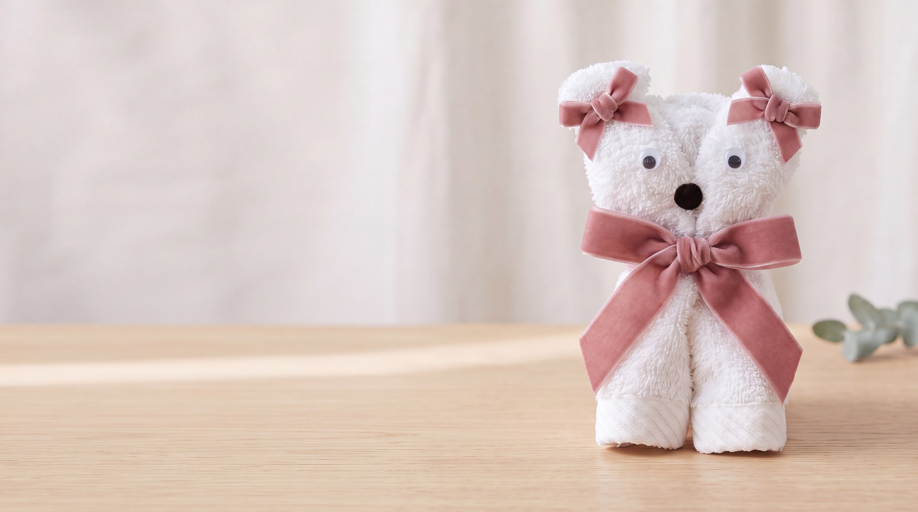Handtuch-Kätzchen aus einem weißen Gästetuch mit Wackelaugen und rosa Samtschleifen auf einem hellen Holztisch